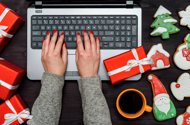 Top view of woman hands typing on open laptop keyboard on table with Cristmas decorations (gift boxes and gingerbread cookies), copy space. Flat lay - Φωτογραφία, εικόνα