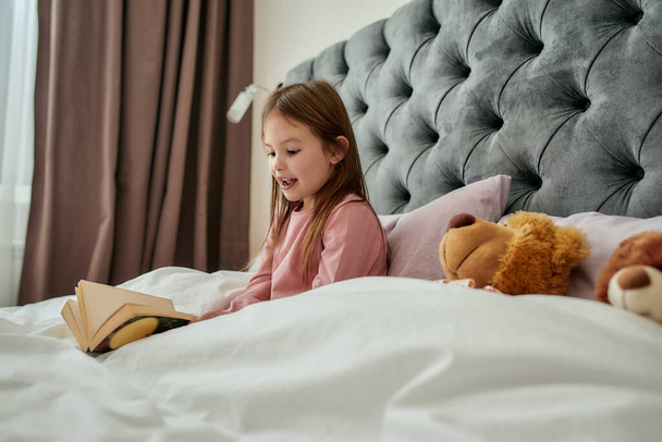 A small girl sitting in a bed wearing pyjamas covered with blanket reading her book and smiling with a teddy bear near to her in a huge bright bedroom at home - Φωτογραφία, εικόνα