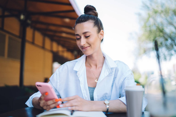 Θετική γυναίκα σε casual outfit κάθεται στο τραπέζι καφέ με σημειωματάριο και takeaway καφέ, ενώ χρησιμοποιώντας smartphone στην καλοκαιρινή βεράντα κατά τη διάρκεια της ημέρας - Φωτογραφία, εικόνα