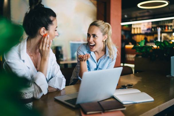 Young cheerful female colleagues in casual wear sitting at table with laptop and having fun together after meeting and discussion in cosy workplace - Foto, Bild
