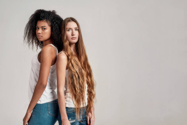 Two young diverse women wearing white shirts looking at camera while standing together isolated over grey background - Foto, Imagem