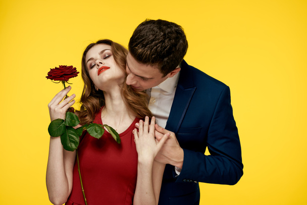 ロマンチックな男は、黄色の背景に彼の手にバラと赤いドレスの女性を抱擁 - 写真・画像