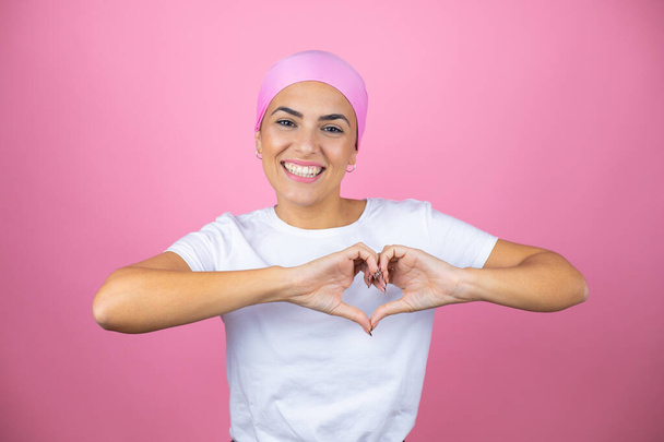 Jovem mulher bonita vestindo lenço rosa sobre fundo rosa isolado sorrindo no amor mostrando símbolo do coração e forma com as mãos - Foto, Imagem