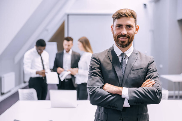 Portrait of a positive looking young business professional standing with his arms crossed with colleagues talking in the background. - Фото, изображение