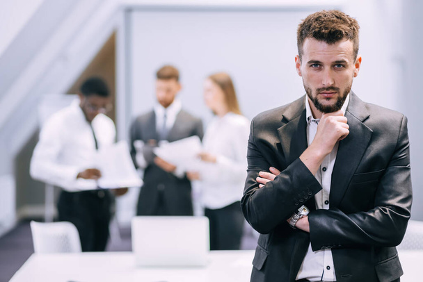 młody biznesmen stojący w nowoczesnym biurze wykazujący się zaufaniem i motywacją w pracy, szczęśliwy millenium europejski mężczyzna CEO pozowanie w miejscu pracy, przywództwo, koncepcja sukcesu. - Zdjęcie, obraz