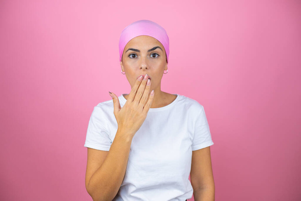 Junge schöne Frau mit rosa Kopftuch auf isoliertem rosa Hintergrund überrascht ihren Mund zu bedecken - Foto, Bild