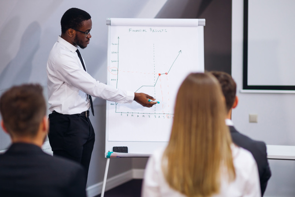 Serious African American mentor giving presentation explains sales growth graph by teaching different trainees in group meeting, new corporate plan on flipchart - Foto, Bild