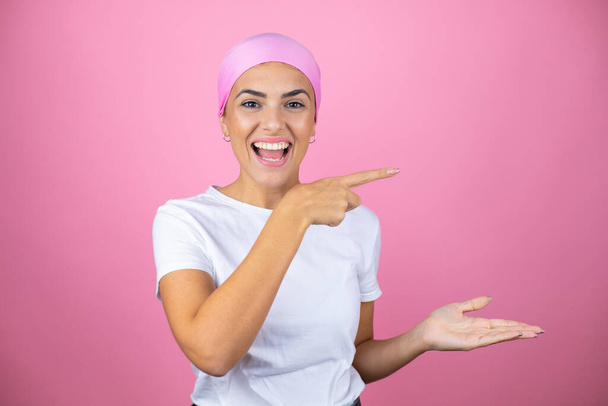 Young beautiful woman wearing pink headscarf over isolated pink background surprised, showing and pointing something that is on her hand - Fotoğraf, Görsel