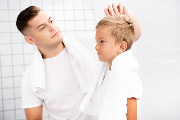 Padre con asciugamano sopra il collo in piedi vicino e fissare i capelli del figlio in bagno su sfondo sfocato  - Foto, immagini