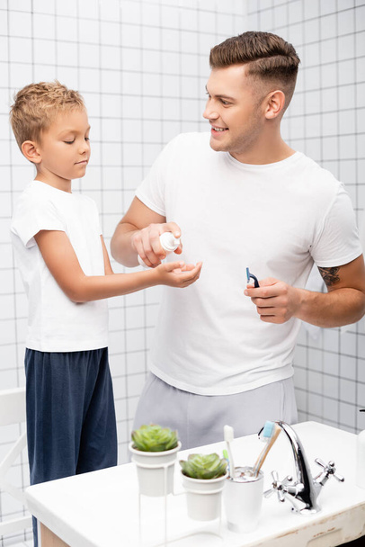 Šťastný otec s bezpečnostní holicí strojek při pohledu na syna při mačkání pěny na holení v blízkosti umyvadla v koupelně - Fotografie, Obrázek
