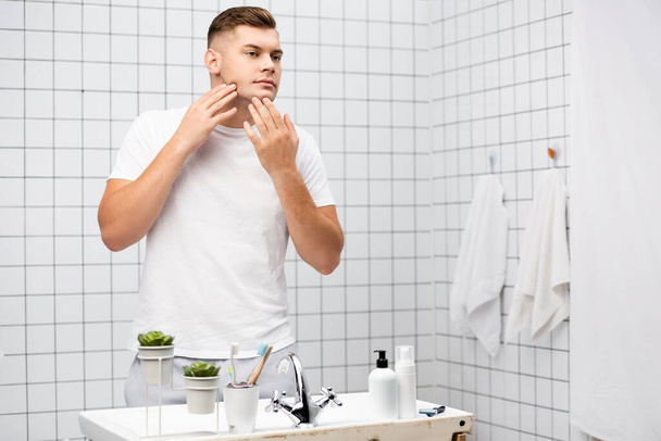 Молодой мужчина трогает лицо, стоя возле раковины с туалетными принадлежностями в ванной комнате - Фото, изображение