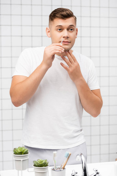 Συμπυκνωμένος νεαρός άνδρας που κρατάει τσιμπιδάκια κοντά στα χείλη στο μπάνιο - Φωτογραφία, εικόνα