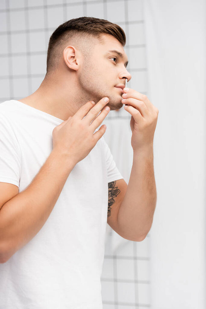 Side view of focused man looking away while plucking hair from nose in bathroom - Photo, Image