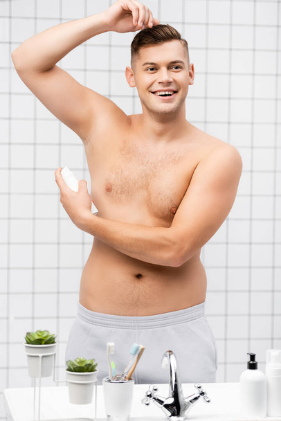 Uomo sorridente senza maglietta con deodorante mentre in piedi vicino al lavandino con articoli da toeletta in bagno - Foto, immagini