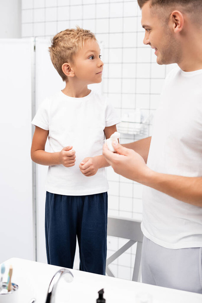 Excited preschooler boy looking at father with dental floss holder while standing on chair in bathroom - Photo, Image