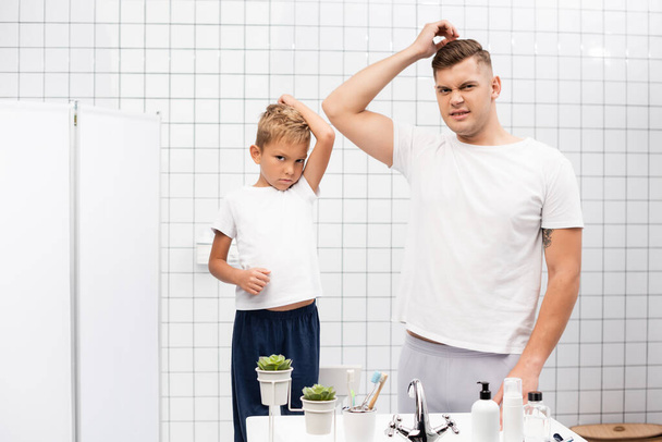 Πατέρας και γιος με μασχάλες μυρίζουν άσχημα κοιτάζοντας κάμερα, ενώ στέκεται κοντά στο νεροχύτη με προϊόντα περιποίησης στο μπάνιο - Φωτογραφία, εικόνα