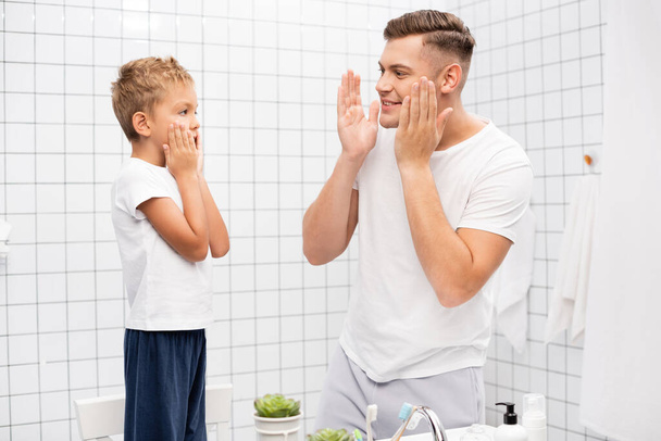 Sorridente padre con le mani vicino guance guardando il figlio applicare lozione da barba mentre in piedi sulla sedia in bagno - Foto, immagini