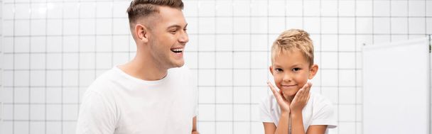 Fröhlicher Vater lacht, während er den Sohn mit den Händen in der Nähe der Wangen im Badezimmer ansieht, Banner - Foto, Bild