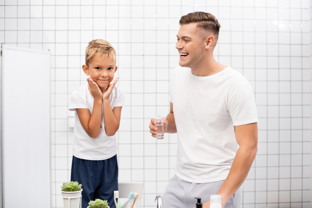 Fröhlicher Vater mit Rasiercreme lacht, während er seinen Sohn mit Händen in der Nähe von Wangen auf einem Stuhl im Badezimmer ansieht - Foto, Bild