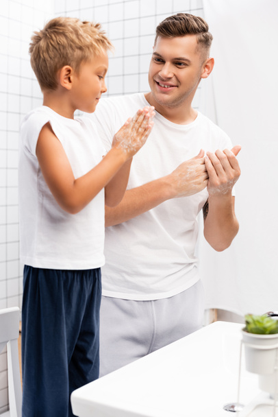 Smiling father looking at son rubbing soap between hands while standing on chair near washbasin in bathroom - Foto, Bild