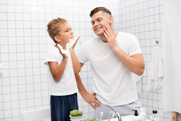 Усміхнений батько з рукою на стегні дивиться на сина, застосовуючи піну для гоління, стоячи на стільці у ванній
 - Фото, зображення