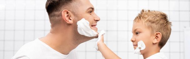 Side view of son applying shaving foam on face of smiling father in bathroom, banner - Fotografie, Obrázek