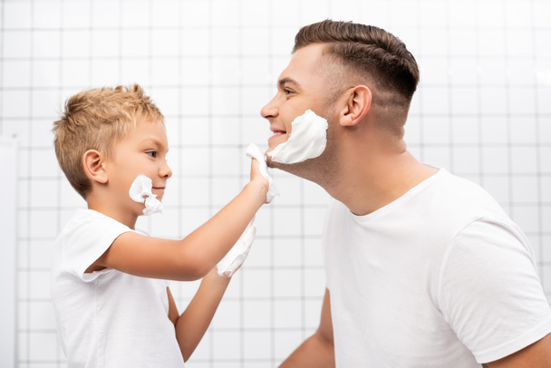 Side view of son applying shaving foam on face of happy father in bathroom - Foto, Bild