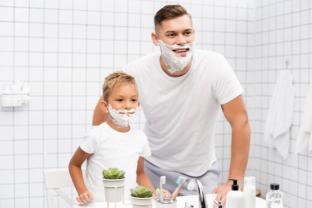 Smiling father and son with shaving foam on faces standing near sink in bathroom - Foto, Imagen