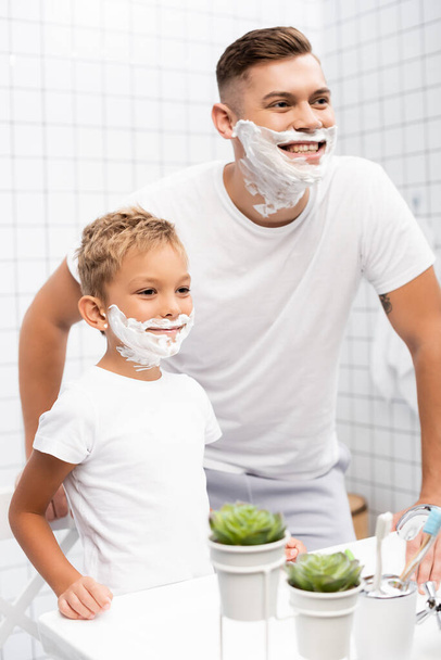 Feliz hijo con espuma de afeitar en la cara de pie cerca de padre sonriente apoyado en el lavabo en el baño - Foto, Imagen