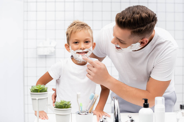 Улыбающийся отец обнимает и трогает подбородок сына с пеной для бритья на лице в ванной - Фото, изображение