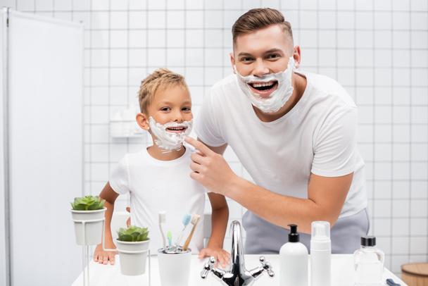 Happy father standing near son with shaving foam on face and touching chin while looking at mirror in bathroom - Foto, Bild