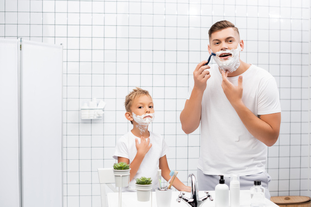 Focused man using safety razor while standing near boy with shaving foam on face in bathroom - Photo, Image