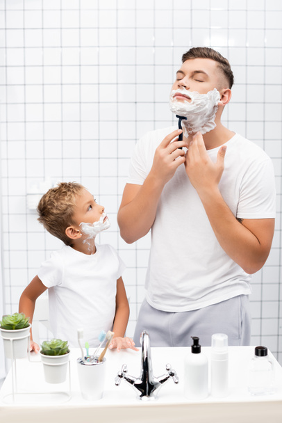 Son with shaving foam on face looking at father with closed eyes using safety razor while standing near sink in bathroom - Foto, Bild