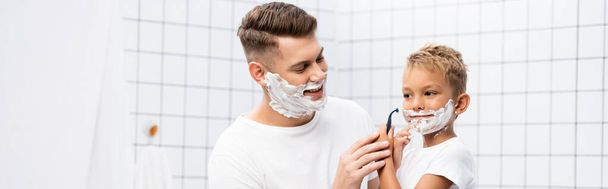 Hymyilevä isä katselee poikaa partavaahto kasvoilla pitäen partaveitsi lähellä poskea kylpyhuoneessa, banneri - Valokuva, kuva