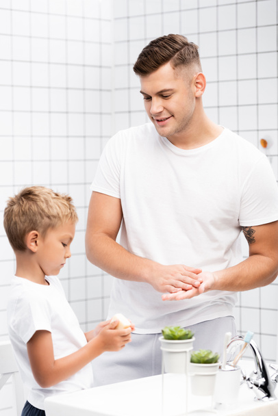 Pozitivní otec mytí rukou a při pohledu na syna s mýdlem stojí v blízkosti umyvadla v koupelně - Fotografie, Obrázek