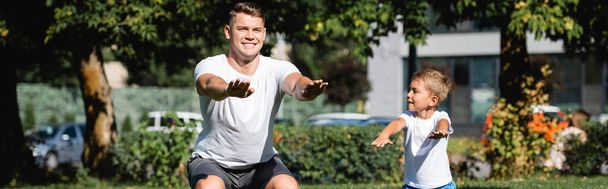 Son with outstretched hands looking at father in sportswear squatting in park on blurred background, banner - Fotografie, Obrázek