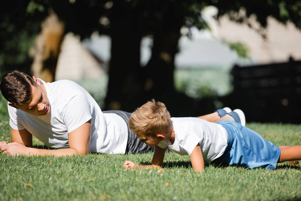 Father and son in sportswear doing plank on grass in park on blurred background - Photo, image
