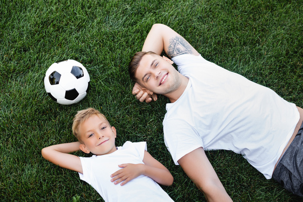 Top view of smiling young adult man and boy with hands behind heads lying near ball on grass  - Foto, imagen