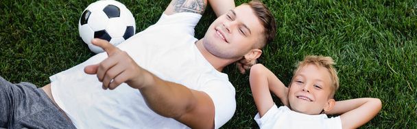 Vista aerea del figlio sorridente che guarda la fotocamera mentre giace vicino al padre che punta con il dito sull'erba, banner - Foto, immagini