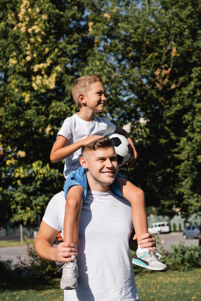 Happy son holding ball and riding piggyback on smiling father in park on blurred background  - Photo, image