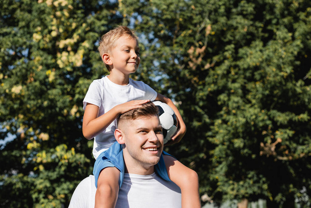 Uśmiechnięty syn z piłką jazda na barana na szczęśliwy ojciec w parku na rozmytym tle  - Zdjęcie, obraz