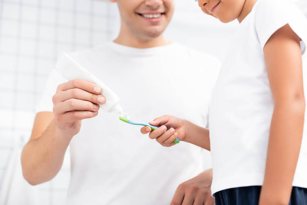Gülümseyen oğlu, babasının yanında duran diş fırçasıyla banyoda diş macununu sıkan bulanık arka planda. - Fotoğraf, Görsel