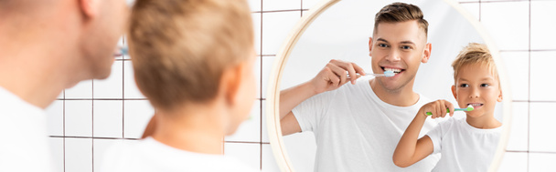 Cheerful father and son brushing teeth while looking at mirror in bathroom on blurred foreground, banner - Фото, зображення