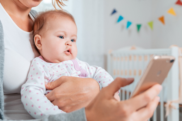 Portrait of a baby sitting in his mother's arms and looking at the smartphone that she uses. Concept of teaching and entertaining children using the Internet. - Foto, imagen