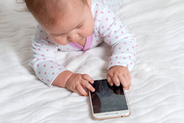 Um bebê em sliders está deitado na cama e brincando com um smartphone. Vista superior. Conceito de jogos infantis com dispositivos modernos. - Foto, Imagem