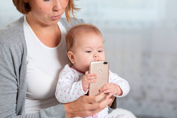 Una madre sostiene al bebé en sus brazos y le da un teléfono inteligente, que está tratando de comer. Concepto de aprendizaje y entretenimiento infantil con tu smartphone. - Foto, imagen