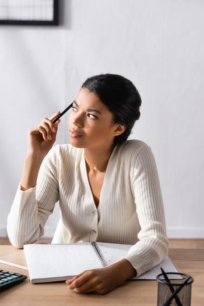 Thoughtful african american woman holding pen and looking away while sitting at desk in office on blurred background - Photo, Image