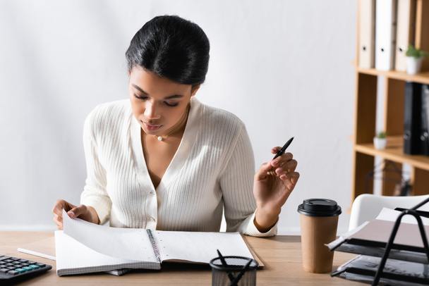 Focused african american woman with pen, looking at blank notebook while sitting at desk in office on blurred background - Fotó, kép