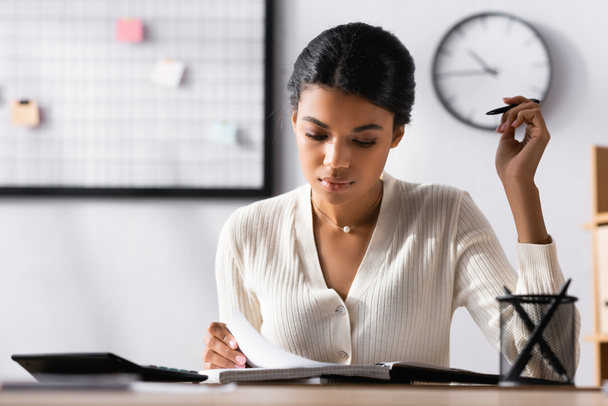 Surface level of focused african american woman looking at notebook while sitting at desk in office on blurred background - Photo, Image