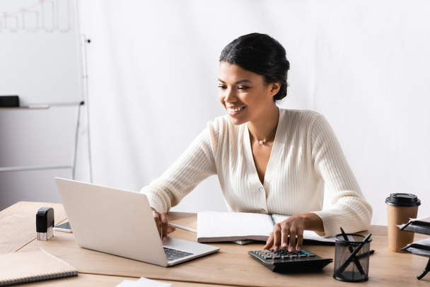 Mutlu Afro-Amerikan kadın bilgisayarına bakarken ofiste hesap makinesinde bulanık arka planda. - Fotoğraf, Görsel